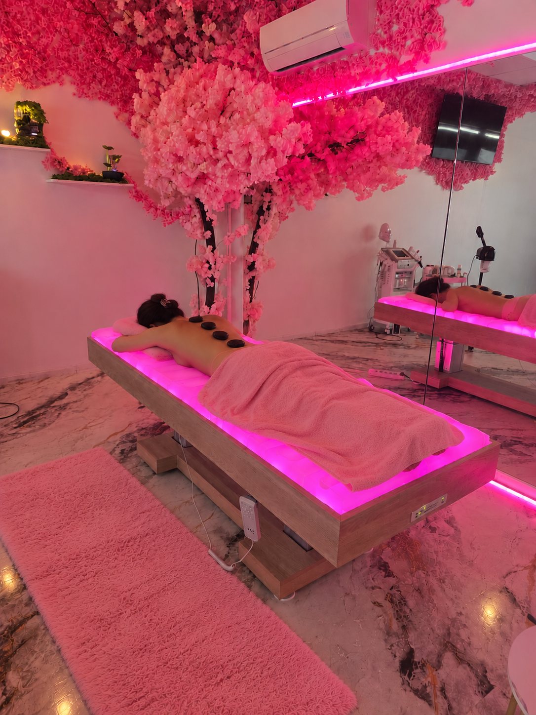 Une cliente détendue sur la table de massage à matelas d’eau éclairée de rose, des pierres posées sur le dos, sous le cerisier en fleurs de l’institut.